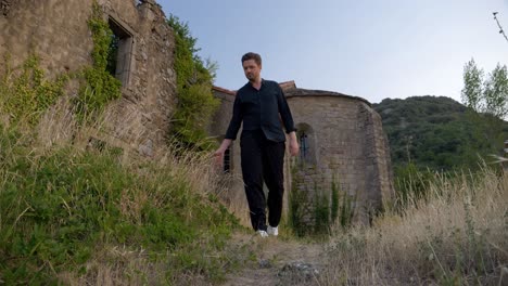 Slowmotion-low-shot-of-a-man-dressed-in-black-walking-on-a-grassfield-outside-some-castle-ruins-during-sunset