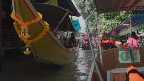 Establishing-shot,-boat-with-tourists-passing-by-and-waving-with-peace-sign-in-Thailand-Floating-Market,-pump-boat-fall-in-line-in-the-background