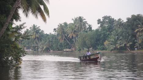 Local-man-on-a-traditional-boat-on-a-river-flowing-near-palm-trees,-Kerala-Backwaters,-India