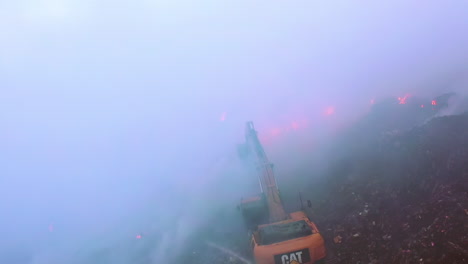 Excavator-fighting-a-wild-fire,-surrounded-by-blue-smoke-rising,-on-a-dark-evening,-in-Amazon,-Brazil,-South-America---Aerial,-drone-shot