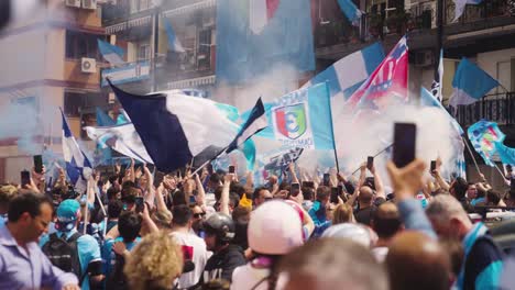 Slow-motion-shot-of-jumping-soccer-fans-celebrating-Championship-of-Naples-outdoors-on-street-in-City-during-sunny-day---waving-flags-and-decorated-balcony-of-house-in-background