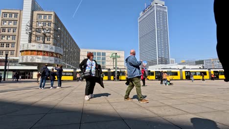 Tourists-Walking-Across-Alexanderplatz-Square-In-Berlin-Taking-Photos-With-Yellow-Tram-Going-Past-In-Background