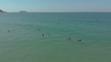 Toma-De-Drone-De-Personas-Surfeando-En-El-Océano-Claro-Y-Cristalino-De-La-Playa-De-Santinho,-Brasil,-Esperando-Las-Olas,-En-Sus-Trajes-De-Surf-En-Un-Día-Soleado