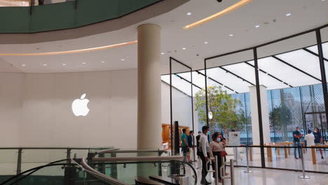 Apple-Store-Dubai-Mall-Septiembre-De-2020-Vista-Panorámica-Con-Un-Gran-Logotipo-Y-Un-Hombre-Con-Mujeres,-Tal-Vez-Un-Aspecto-De-Negocios-Asiático-Vestido-Con-Máscaras-Médicas-Pasando-Frente-A-La-Entrada-Sosteniendo-Teléfonos-Inteligentes