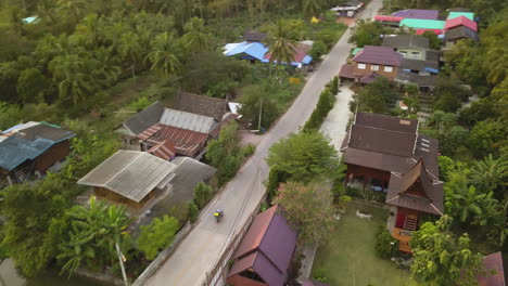 Motobike-Riders-on-Thailand-Countryside-Road-Aerial,-Tropical-Vegetation-and-Authentic-Houses