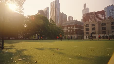 Wide-shot-of-two-teams-playing-soccer-on-grass-field-surrounded-by-skyscraper-in-NYC-at-sunset