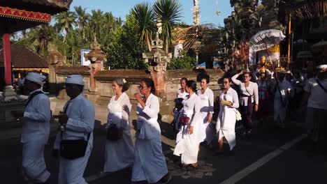 Priests-Playing-Bells-in-Walking-Procession-of-Balinese-Hinduism-Traditional-Ceremony-wearing-White-Clothes,-Women-carry-Golden-Offerings-on-their-heads,-Bali-Indonesia