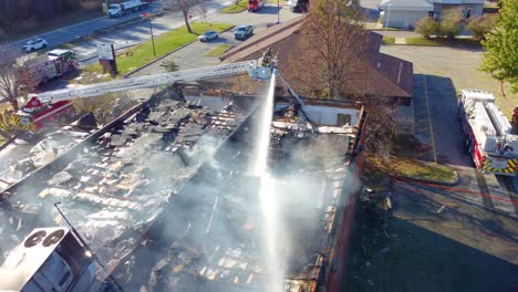 Two-Hoses-Simultaneously-Putting-Out-Smoking-Roof-of-Large-Building,-Wide-Aerial