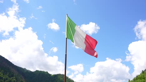 Italienische-Flagge-Vor-Blauem-Himmel-Mit-Weißen-Wolken,-Berge-Im-Hintergrund