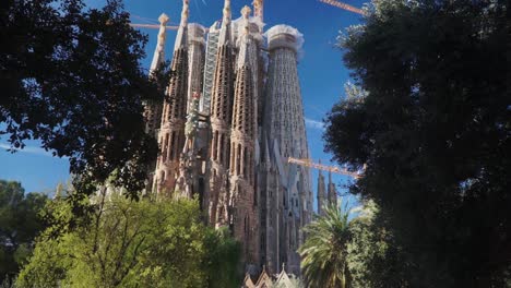 Toma-En-Movimiento-Lento,-árboles-Que-Revelan-La-Vista-Panorámica-De-La-Iglesia-De-La-Sagrada-Familia-En-Barcelona,-España,-Pájaro-Volando-En-El-Fondo