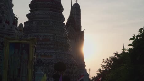 Toma-Panorámica-Hacia-La-Izquierda,-árbol-Que-Revela-Una-Vista-Panorámica-Del-Templo-En-Bangkok,-Tailandia,-Banderas-Ondeando-Y-Puesta-De-Sol-En-El-Fondo