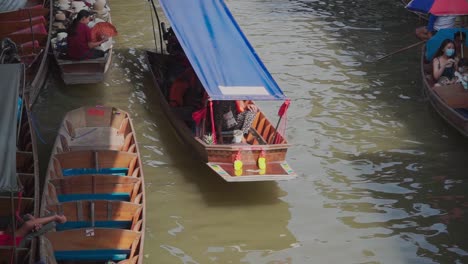 Tiro-Hacia-Abajo,-Barco-Con-Lona-Azul-Pasando-Por-El-Mercado-Flotante-De-Tailandia,-Vendedor-De-Sombreros-Estacionado-A-Un-Lado