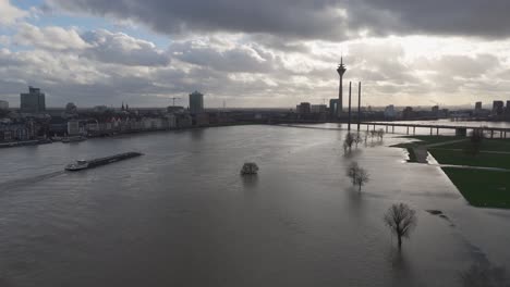 A-barge-sails-up-flooded-Rhine-river,-past-partially-submerged-trees,-Dusseldorf-skyline,drone
