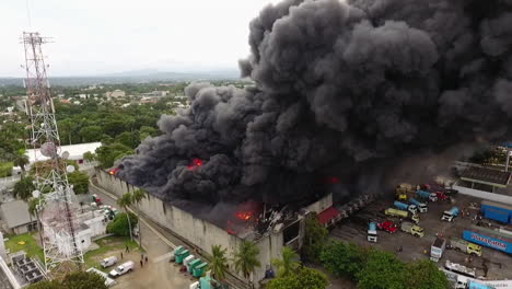 Aerial-view-of-firetrucks-and-firemen-fighting-a-warehouse-in-flames,-black-smoke-rising,-during-day-time,-in-Dominican-republic---pull-back,-drone-shot