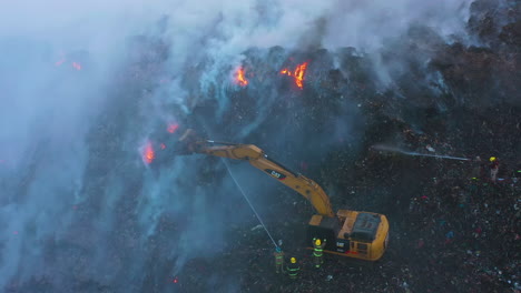 Excavator-and-firemen-trying-to-extinguish-a-wildfire,-blue-smoke-rising,-on-a-dark-evening,-in-Amazon,-Brazil,-South-America---Aerial,-descending,-drone-shot