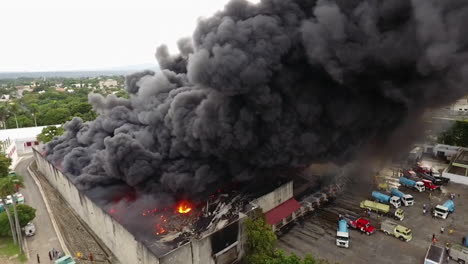 Aerial,-pan,-drone-shot-around-firetrucks-and-firemen-extinguishing-a-warehouse-on-fire,-during-day-time,-in-Dominican-republic