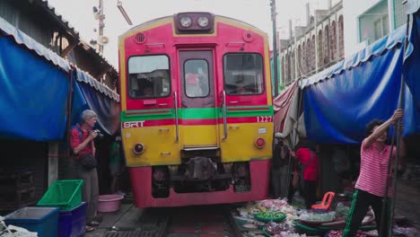 POV-shot,-Train-is-moving-along-the-railroad-of-Maeklong-Market-in-Thailand,-Tourist-takes-picture,-vendors-preparing-their-shops-in-the-background