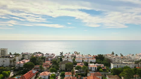 Drone-footage-captures-scenic-vistas-of-coastal-homes-by-beach,-offering-breathtaking-views-of-vast-sea-on-a-sunny-day