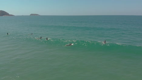 Drone-shot-of-people-surfing-in-the-clear-and-crystal-ocean-of-the-Santinho-Beach,-Brazil,-waiting-for-the-waves,-in-their-surfing-wetsuits-on-a-sunny-day