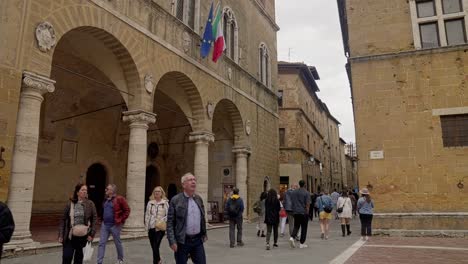 Clock-Tower-At-Palazzo-Comunale-In-Piazza-Pio-II-With-Locals-On-The-Street-Corner-In-Pienza,-Tuscany,-Italy