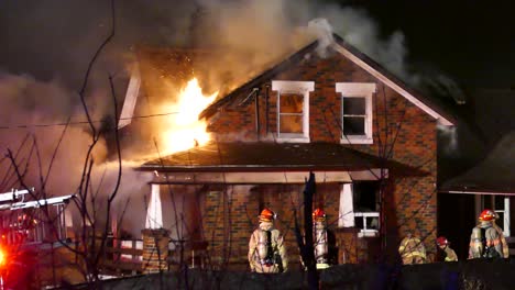 Firefighters-stand-around-fighting-blaze-house-home-fire,-bright-flames-exit-through-side-windows