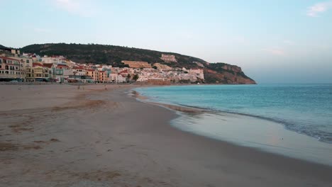 Portugal,-Sesimbra,-Atlantic-Ocean-washes-the-shore-of-the-long-amazing-sandy-beach-with-the-coastal-townscape-in-the-background-after-sunset