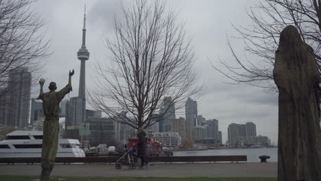 Woman-Walking-With-Stroller,-Downtown-Toronto-City-Skyline-In-Background