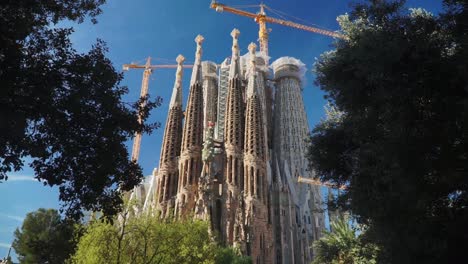 Truck-Shot,-Bright-blue-sky,-trees-revealing-the-ongoing-construction-of-Sagrada-familia-Church-in-the-background