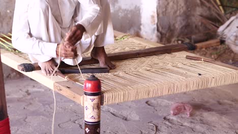 Male-Labourer-Sitting-On-Charpai-Weaving-Thread-Across-Inside-Workshop-In-Sindh,-Pakistan