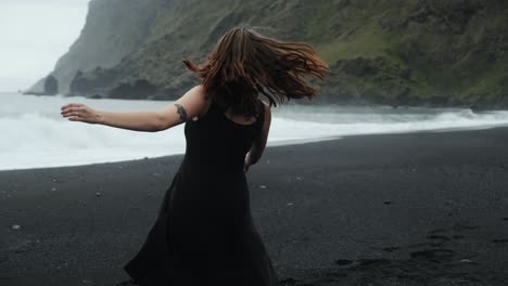 Young-redhead-woman-in-black-dress-dancing-on-black-sand-beach-Iceland,-slow-motion-spinning,-dramatic-waves-seascape