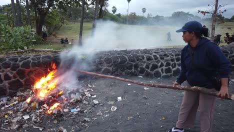 Frau-Mit-Einem-Stock-Verbrennt-Plastikmüll-Und-Erzeugt-Rauch-Und-Feuer,-Asiatischer-Strand-Auf-Bali,-Indonesien,-Giftige-Dämpfe,-Umweltverschmutzung