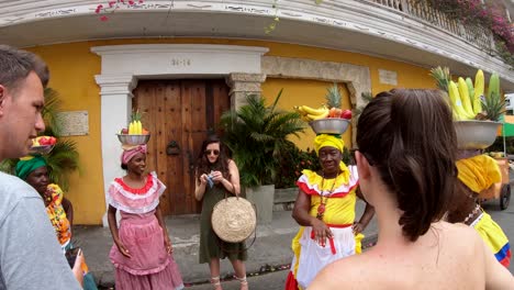 Several-palenqueras-with-colorful-dresses-who-are-balancing-fruit-bowls-over-their-heads-while-speaking-with-tourists-in-the-old-town-of-Cartagena-de-Indias,-Colombia
