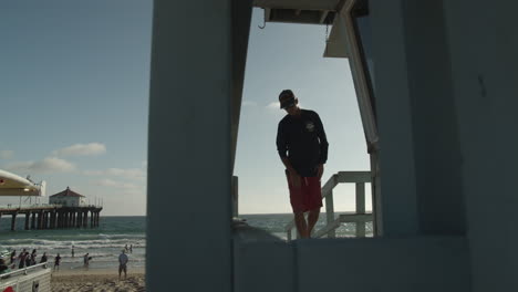 Cinematic-shot-of-lifeguard-walking-to-guard-tower-on-the-beach-with-historic-Manhattan-Beach-pier-and-tourists-in-the-background