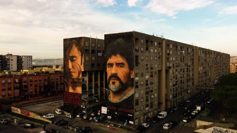 Aerial-orbiting-shot-showing-ESSERI-UMANI-and-Diego-Maradona-Painting-on-Wall-of-apartment-block-lighting-by-sun-during-sunset---Naples-City,-Italy