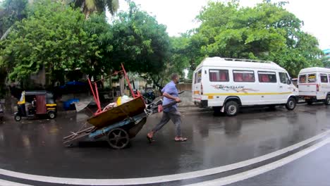 Männlicher-Arbeiter-Mit-Gesichtsmaske-Zieht-Einen-Wagen-Voller-Dinge-Auf-Der-Straße-In-Mumbai,-Indien,-Unter-Dem-Regen---Fischaugenansicht---Zeitlupe