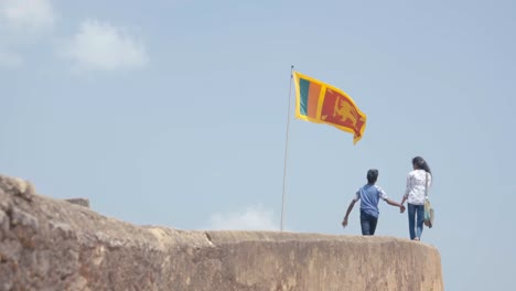 People-Strolling-At-The-Galle-Dutch-Fort-In-Weligama,-Sri-Lanka-With-Sri-Lankan-Flag-Waving-On-A-Sunny-Day---low-angle-slowmo-shot