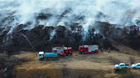 Aerial,-drone-shot-over-firetrucks-and-firemen-fighting-a-wildfire,-in-smoky-nature-smoke-rising,-on-a-cloudy-day,-in-Santo-Domingo,-Dominican-Republic