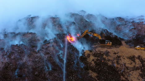 Aerial,-tracking,-drone-shot-of-a-digger-fighting-a-wildfire,surrounded-by-smoke-and-flames,-gloomy-evening,-in-Los-Angeles,-California,-USA