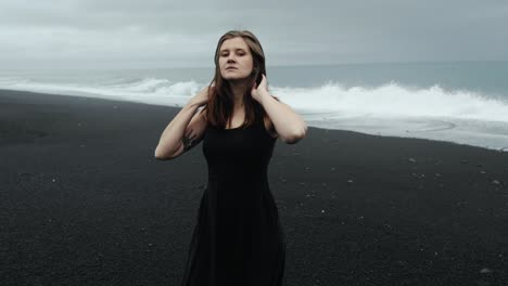 Young-beautiful-woman-in-black-dress-dancing-on-black-sand-beach-Iceland,-slow-motion-walking,-dramatic-waves-seascape
