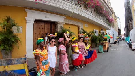 Touristen-Posieren-Neben-Palenqueras-Für-Ein-Foto,-Während-Sie-In-Einer-Straße-Der-Altstadt-Von-Cartagena-De-Indias,-Kolumbien,-Obstschalen-über-Ihren-Köpfen-Balancieren
