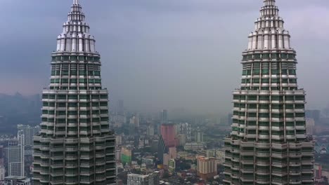 Cinematic-drone-fly-pedestal-up-along-the-iconic-landmark,-Petronas-KLCC-Twins-towers,-capturing-downtown-cityscape-at-central-district,-Kuala-Lumpur-on-a-foggy-day