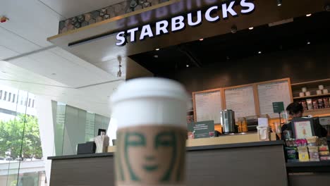 Im-Hintergrund-Ist-Die-Amerikanische-Multinationale-Kaffeemarke-Starbucks-Coffee-Store-Zu-Sehen,-Während-Im-Vordergrund-Ein-Kaffee-Pappbecher-Steht