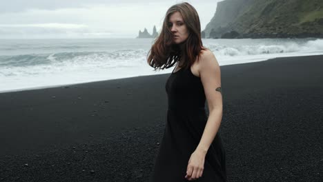 Young-beautiful-woman-in-black-dress-dancing-on-black-sand-beach-Iceland,-slow-motion-walking,-looking-into-camera