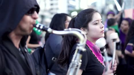 Ein-Mann-Mit-Sonnenbrille-Spielt-Saxophon-Und-Eine-Gruppe-Frauen-Spielt-Schlagzeug-Bei-Einem-Marsch-Und-Protest-Während-Des-Internationalen-Frauentags-In-Quito