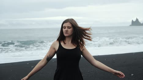 Young-beautiful-woman-in-black-dress-dancing-on-black-sand-beach-Iceland,-dramatic-waves-seascape,-slow-motion-spinning