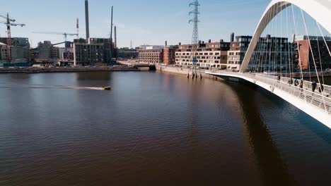 Aerial-view-of-a-boat-driving-under-the-a-white-bridge,-while-people-walk-on-it,-sunny-day,-in-the-Kalasatama-area,-Helsinki-city,-Uusimaa,-Finland---dolly,-drone-shot