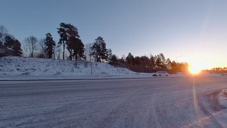 The-snow-covered-road-stretches-ahead,-illuminated-by-the-warm-glow-of-the-setting-sun,-as-cars-pass-by-amidst-the-wintry-landscape,-with-road-signs-guiding-their-way