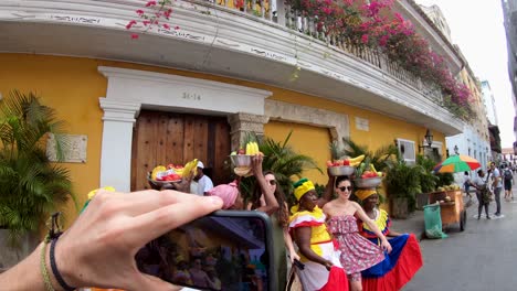 A-picture-is-being-taken-by-a-phone-camera-of-a-group-of-tourists-alongside-palenqueras-as-they-balance-fruit-bowls-on-their-heads-on-a-street-of-the-old-town-of-Cartagena-de-Indias,-Colombia