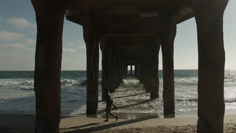 Cinematic-shot-of-man-walking-underneath-beach-pier-in-slow-motion-in-slow-motion
