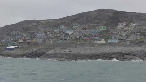 Panoramablick-Auf-Die-Küste-Grönlands-Mit-Häusern-Auf-Einem-Hügel-Und-Wellen,-Die-Auf-Felsen-Schlagen